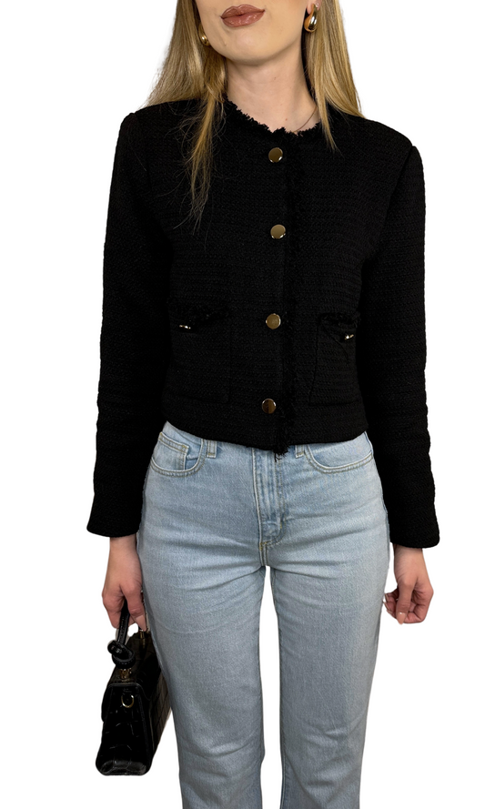 A woman wearing a tweed jacket with a mock neckline, long sleeves, and button-down closure. The jacket is black with gold buttons, and she is also wearing light blue jeans and carrying a black bag.