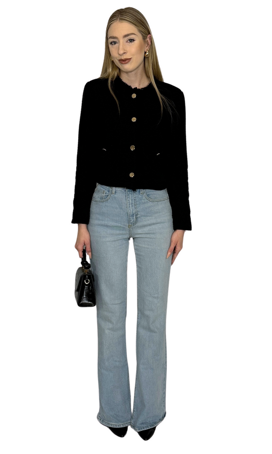 A woman wearing a tweed jacket with a mock neckline, long sleeves, and button-down closure. The jacket is black with gold buttons, and she is also wearing light blue jeans and carrying a black bag.